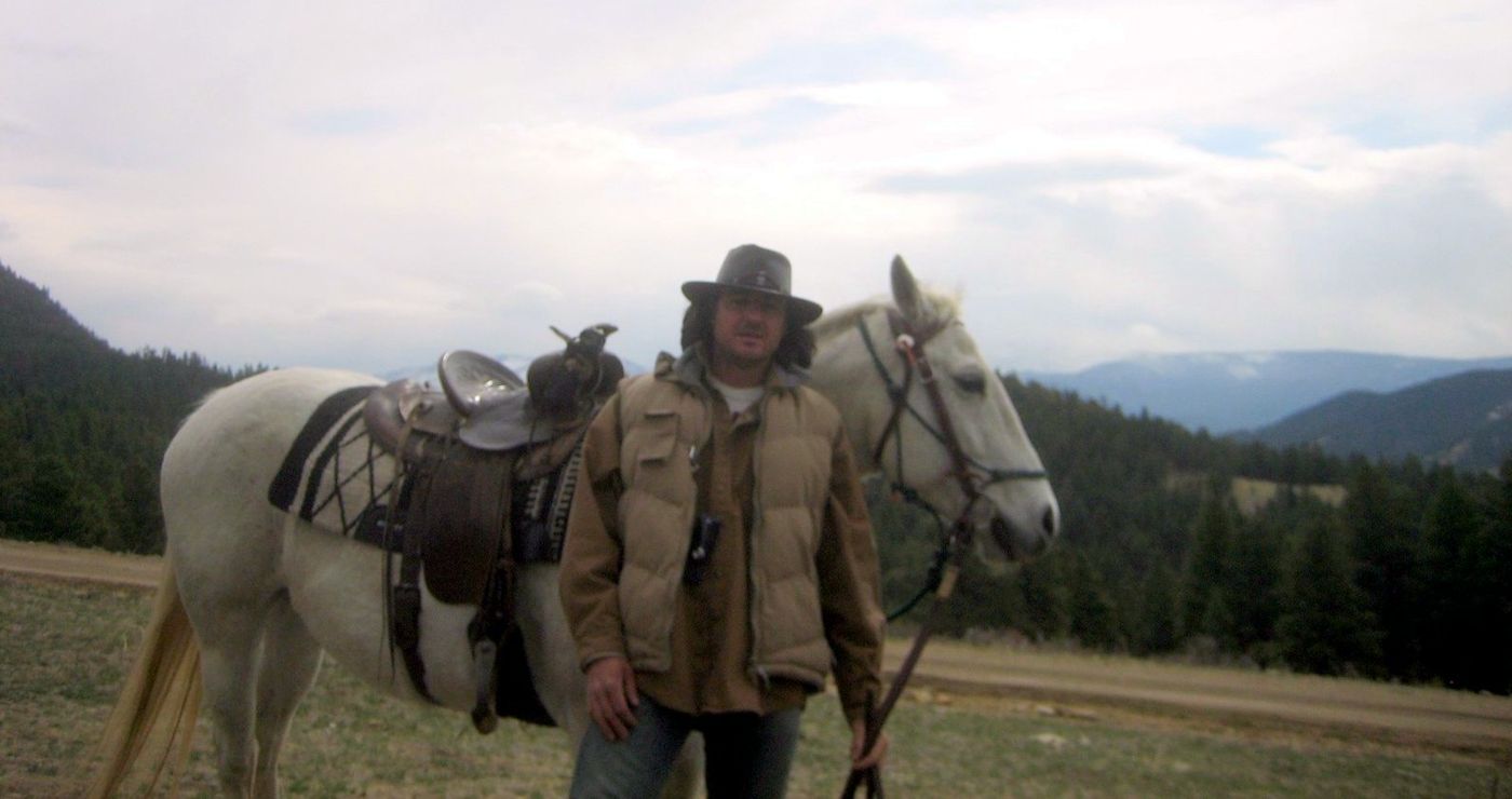 Le guide en chapeau de cowboy à côté d'un cheval blanc sellé, montagnes en arrière-plan